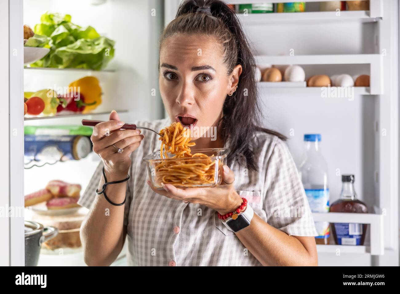 Verängstigte hungrige Frau im Schlafanzug isst nachts Spaghetti im Kühlschrank. Stockfoto