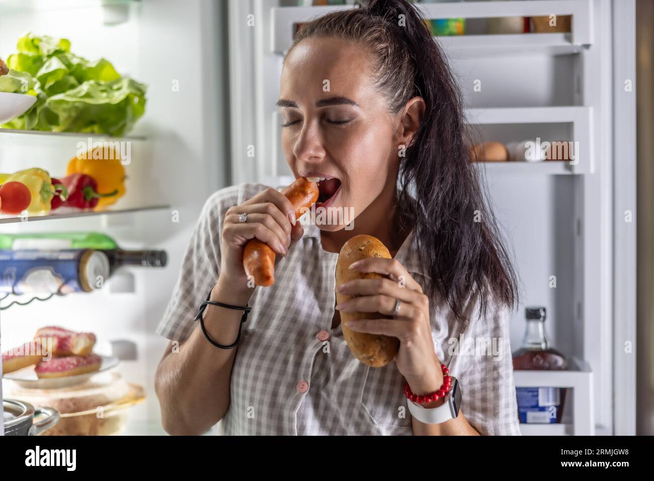 Hungrige Frau im Pyjama isst abends am Kühlschrank Würstchen mit Brot. Stockfoto
