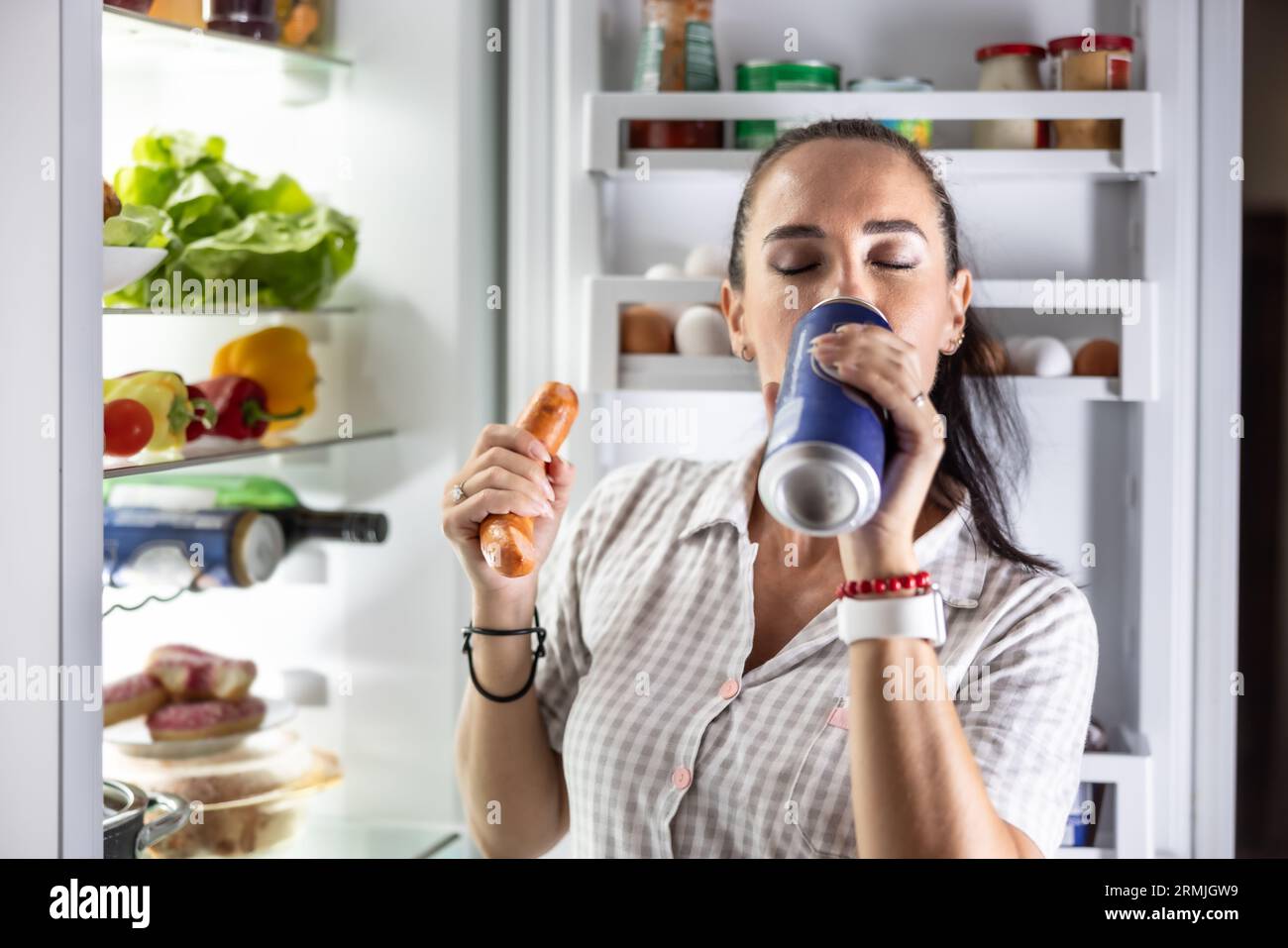 Hungrige Frau im Pyjama trinkt Bier und isst abends Würstchen am Kühlschrank. Stockfoto
