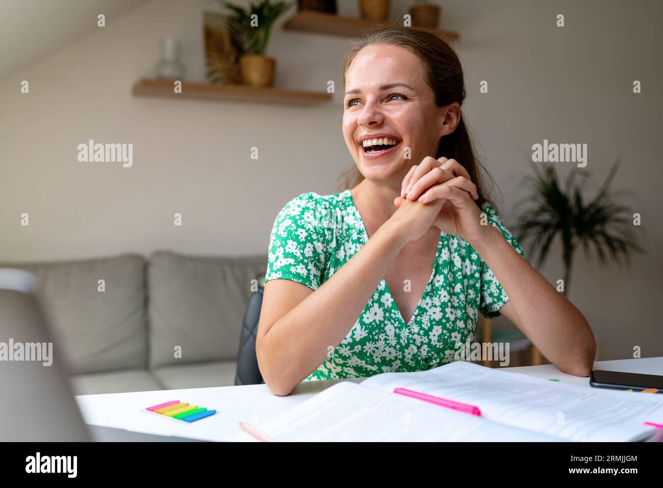 Glückliche junge Erwachsene elegante Frau in grünem Kleid sitzt zu Hause am Tisch und lacht. Stockfoto