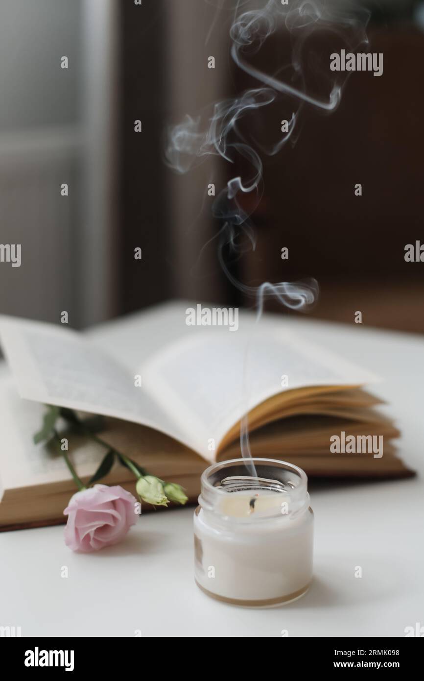 Stillleben-Details im Wohnzimmerinneren. Offenes Buch mit Blume, brennende Kerze mit Rauch. Lesen und ausruhen. Gemütliches Zuhause Stockfoto