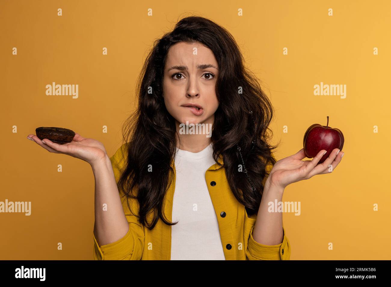 Verwirrte dunkelhaarige, süße Dame, die während des Fotoshootings zwischen einem kalorienreichen Keks und einer kalorienarmen Frucht wählt. Schwieriges Auswahlkonzept Stockfoto