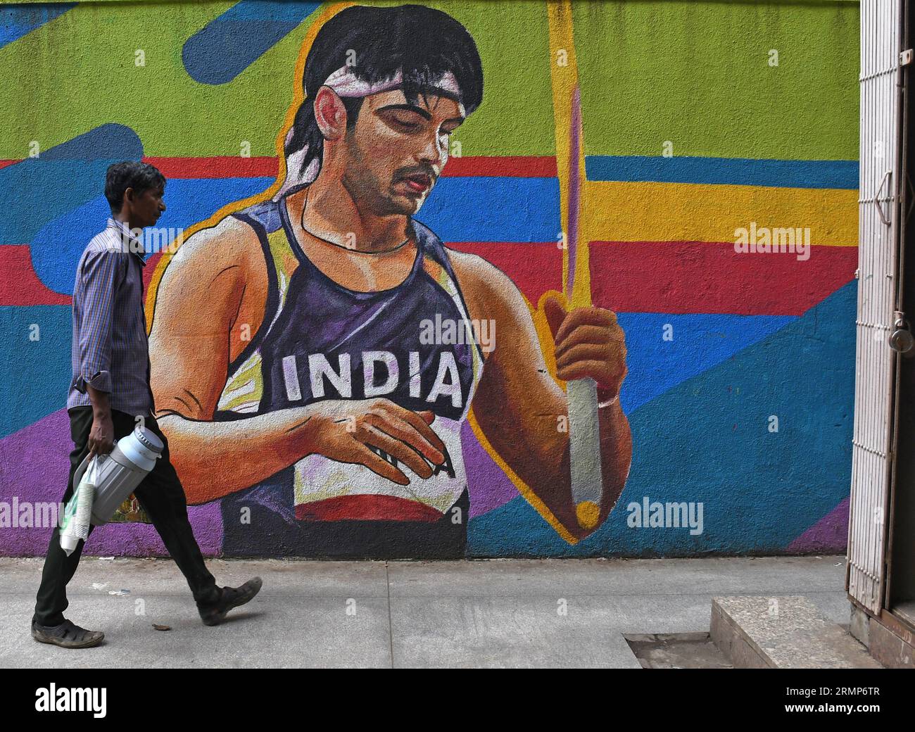 Ein Teehändler mit einer Plastikthermos geht an einem Wandgemälde des indischen Leichtathleten Neeraj Chopra in Mumbai vorbei. Neeraj Chopra gewann als erster Indianer die Goldmedaille bei der Leichtathletik-Weltmeisterschaft in Budapest. Er schaffte es, den Speer in einer Entfernung von 88 m zu werfen, was ihm die Goldmedaille vor dem pakistanischen Arshad Nadeem einbrachte, der eine Silbermedaille gewann. Stockfoto