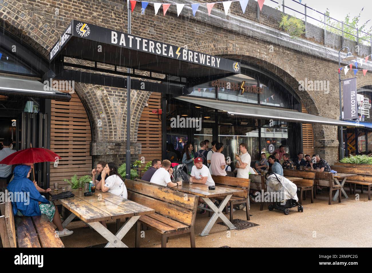 Menschen sitzen und trinken draußen im Regen vor der Battersea Brewery, die sich unter den Bögen im Circus West Village auf der Arches Lane befindet. London, Großbritannien Stockfoto