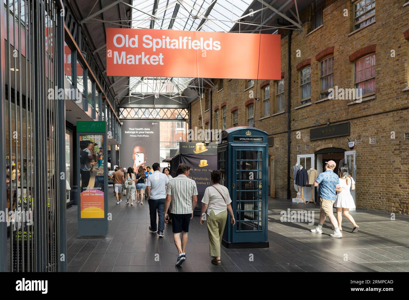 Leute, die durch den überdachten Old Spitalfields Markt gehen, mit einem großen Werbeschild oben und einer grünen Telefonbox. London, England Stockfoto