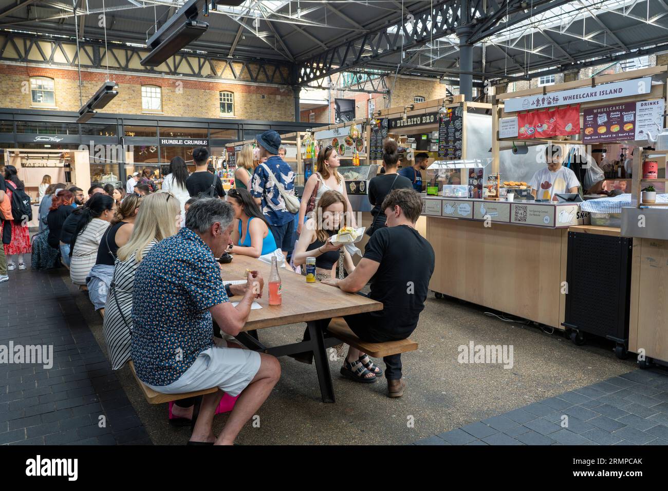 Die Leute, die sich zum Essen und Trinken an Straßenrestaurants und Imbissständen zum Mitnehmen auf dem überdachten Markt der alten Spitalfields hinsetzen. Spitalfields, London, Großbritannien Stockfoto