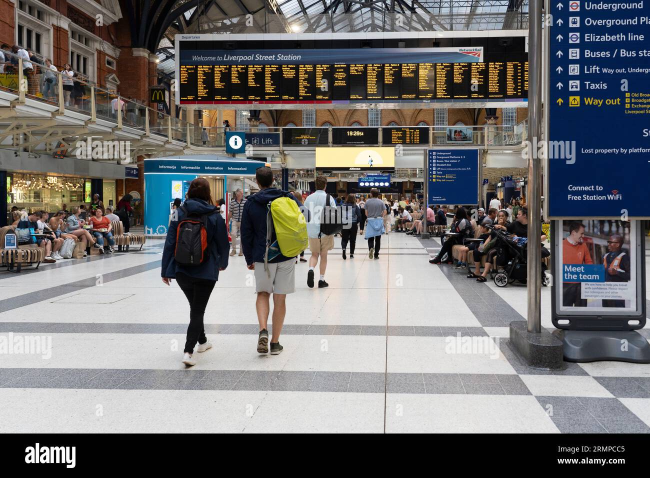 Bahnpassagiere, die durch den Bahnhof Liverpool Street gehen, mit einem Willkommensschild und einer elektronischen Tafel mit den Abfahrtszeiten des Zuges. London, England Stockfoto