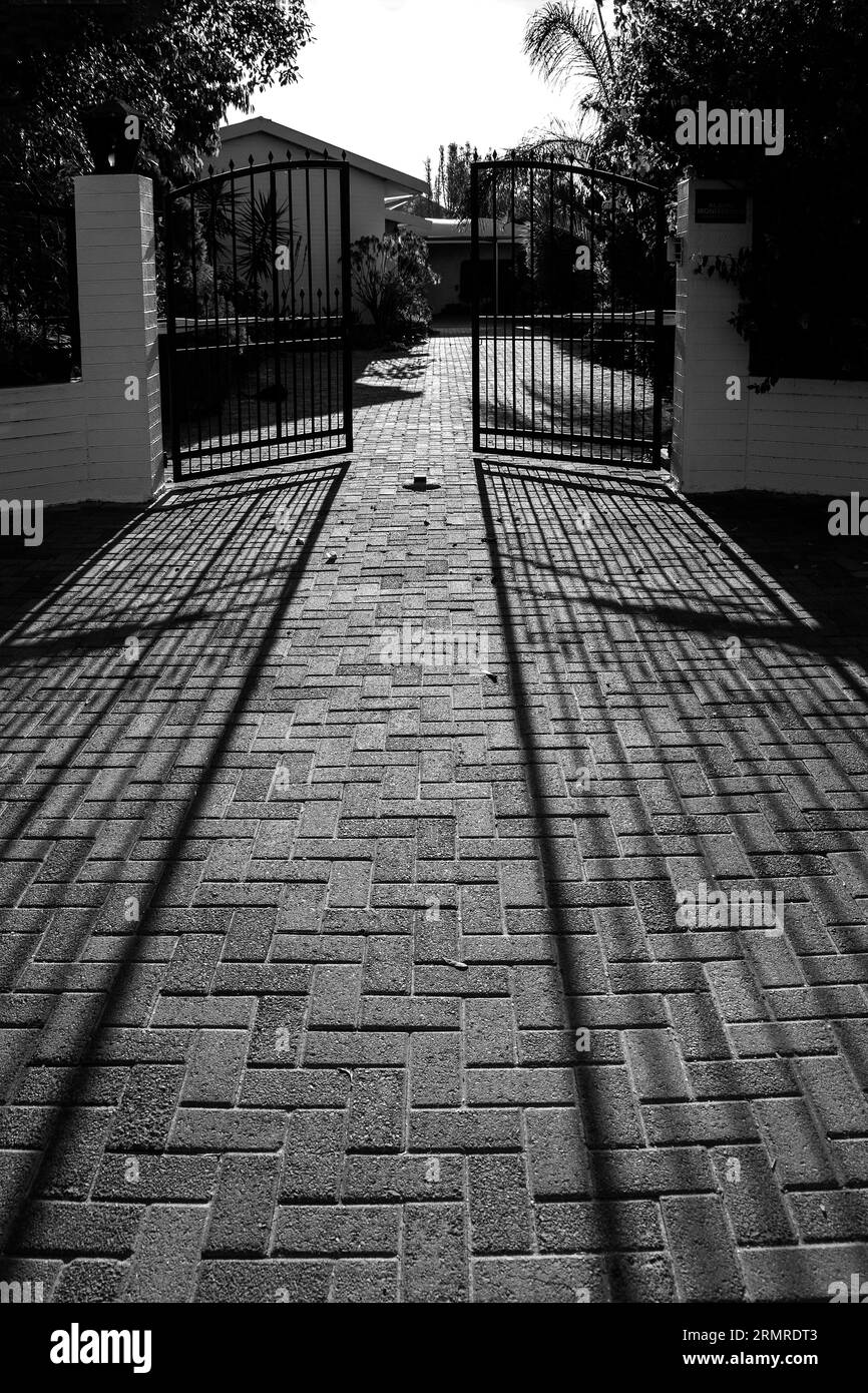 Die Schatten von einem Paar halb offener, schwarzer, finsterer schmiedeeiserner Tore, die sich zur Kamera mit einem weißen Haus dahinter erstrecken. Stockfoto