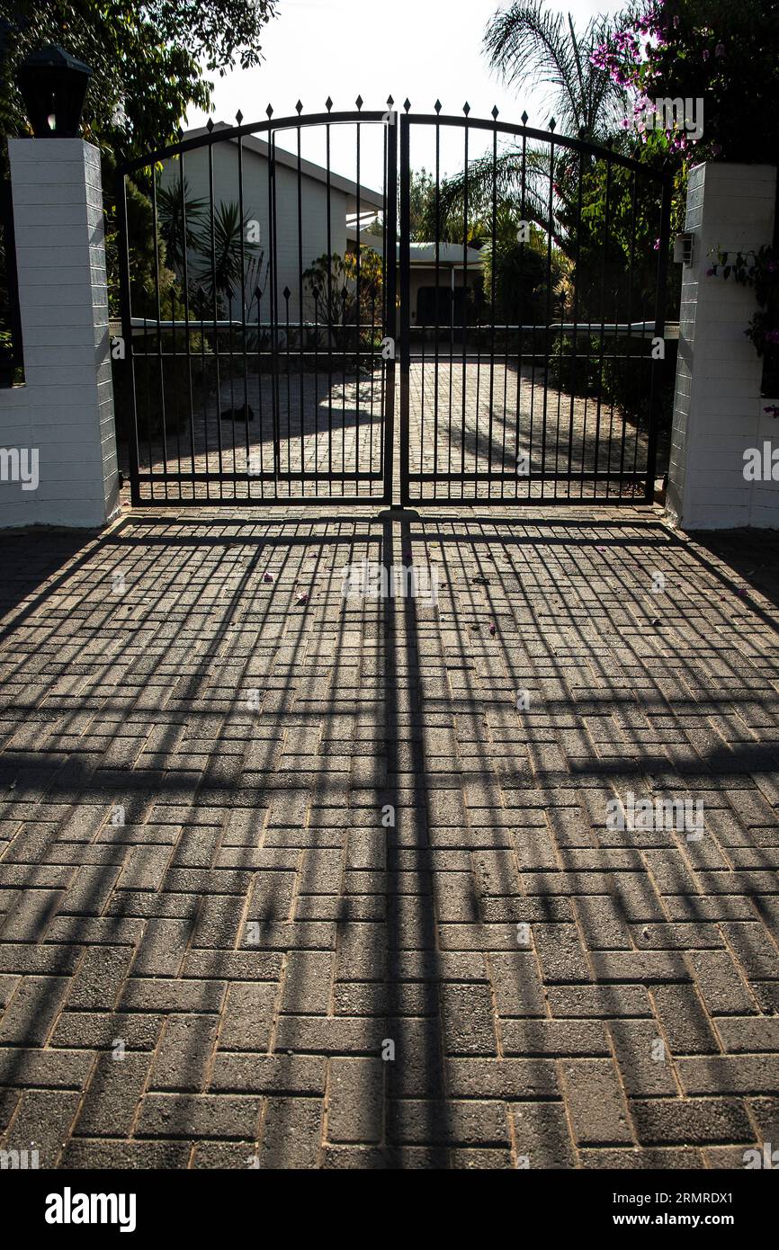 Die Schatten von einem Paar geschlossener, schwarzer, unheimlicher schmiedeeiserner Tore, die sich zur Kamera mit einem weißen Haus dahinter erstrecken. Stockfoto