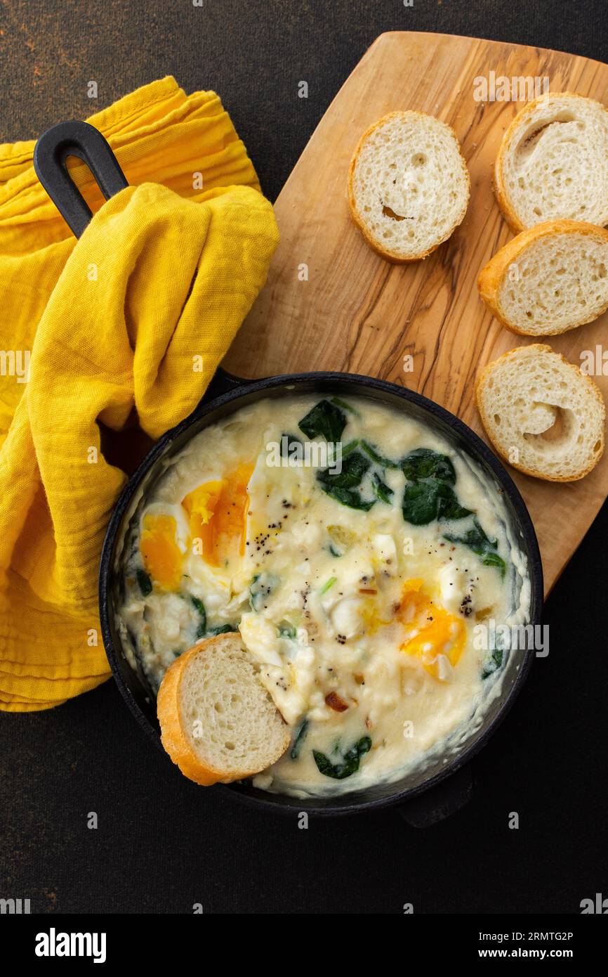 Gesundes Frühstück oder Abendessen, Spiegeleier mit Käse, Spinatblätter und Kokosmilch in einer gusseisernen Pfanne, Baguette auf einem Schneidebrett Stockfoto