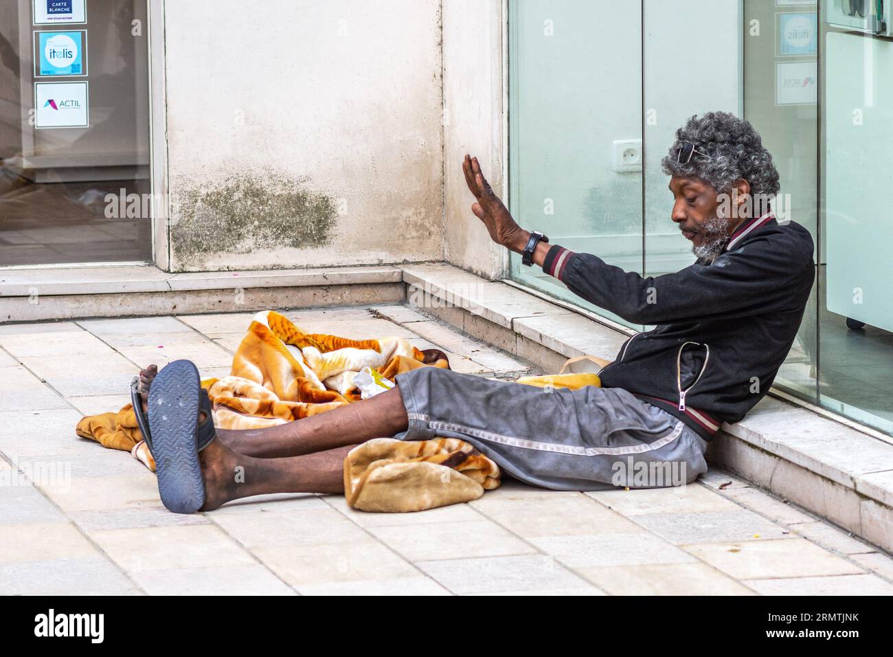 Heimatloser schwarzer Mann, der mit erhobener Hand auf dem Bürgersteig sitzt - Tours, Indre-et-Loire (37), Frankreich. Stockfoto