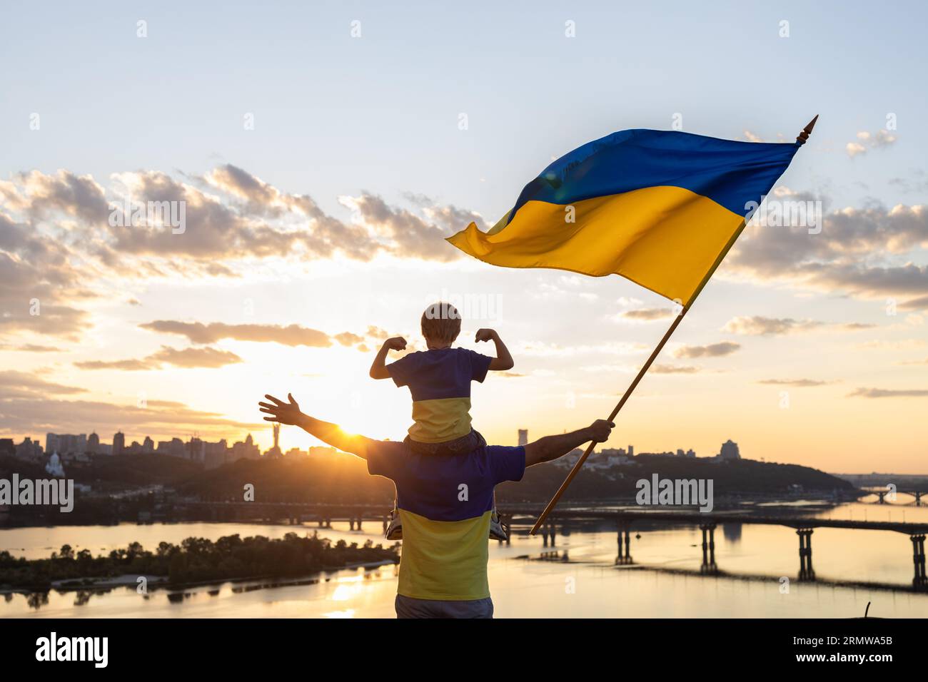 Vater mit Sohn, der auf Schultern sitzt, mit großer Flagge der Ukraine auf dem Dach des Hauses auf dem Ufer des Dnieper Flusses und Landschaft von Kiew. Patriotische Erziehung Stockfoto