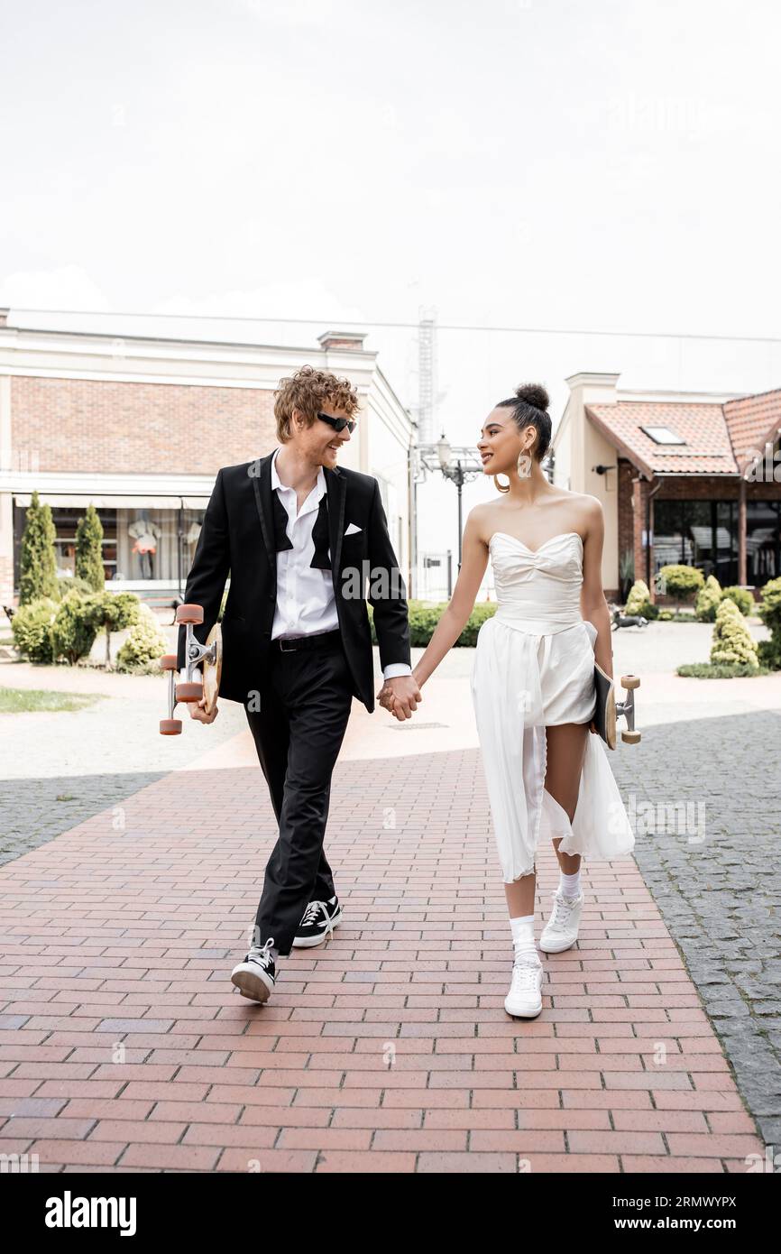 Stilvolles interrassisches Paar mit Longboard- und Skateboardhalterung und Spaziergang in der Stadt, Hochzeitstag Stockfoto