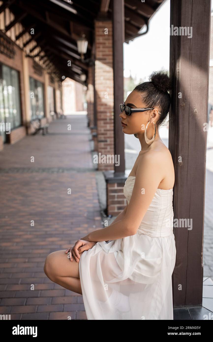 Seitenansicht der bezaubernden afroamerikanischen Frau in Hochzeitskleid und Sonnenbrille auf der urbanen Straße Stockfoto