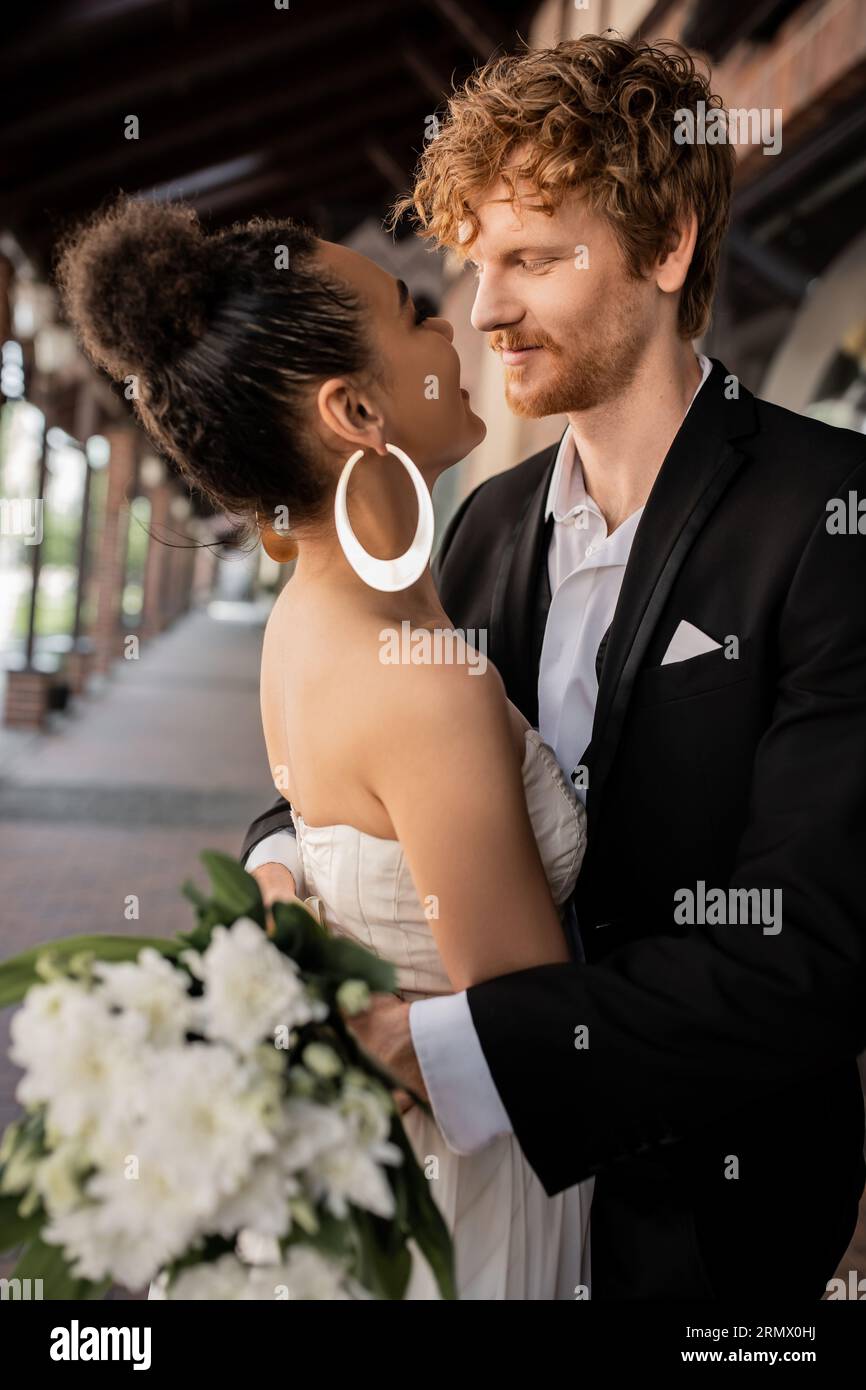 Fröhliches, interrassisches Paar, das sich umgibt und anschaut, Hochzeitskleidung, städtisches Setting Stockfoto