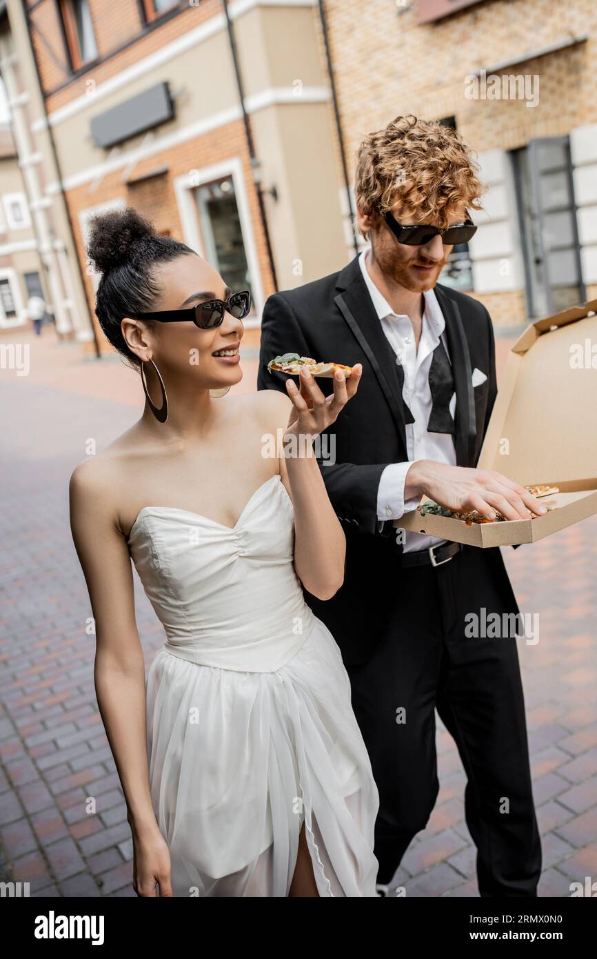 Outdoor-Feier, interrassische Paare mit Pizza in der Stadt, Hochzeitskleidung, Sonnenbrille Stockfoto