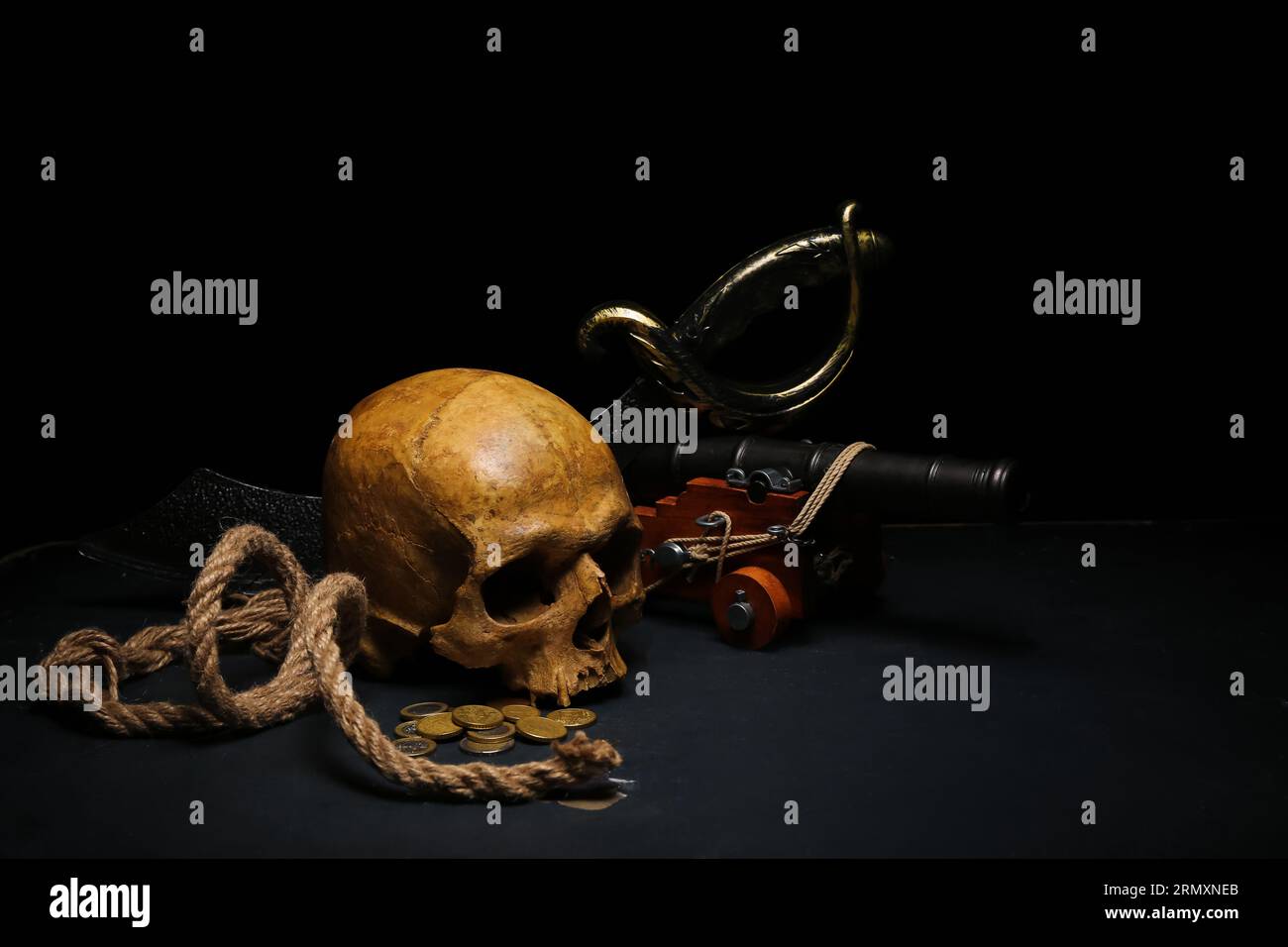 Menschlicher Schädel mit Schwert, Spielzeugmodell der Kanone, Piratenhut und Münzen auf schwarzem Hintergrund Stockfoto