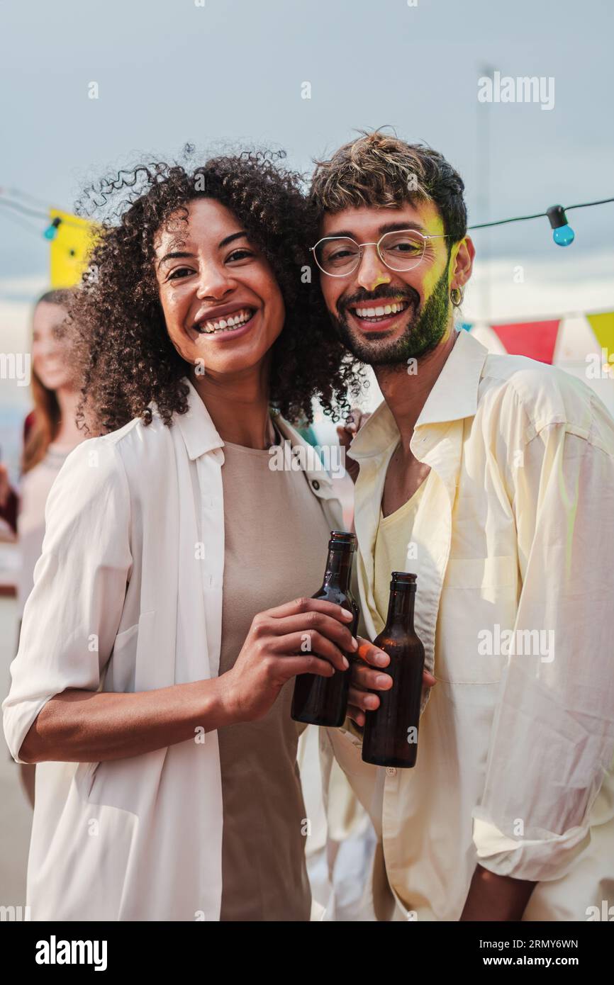 Vertikales Porträt eines jungen erwachsenen, multirassischen Paares, das lächelt, lacht und die Kamera mit Bierflaschen betrachtet. Zwei Leute starren mit ihren besten Freunden auf eine Party. Hochwertige Fotos Stockfoto