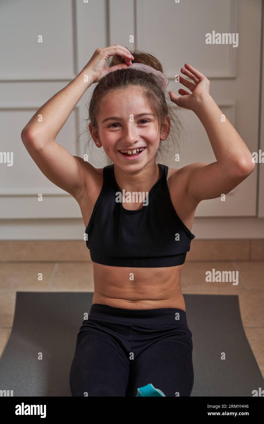 Lächelndes junges Mädchen in Sportkleidung, das sich auf einer grauen Matte ruht und die Kamera anschaut, während es während der Pause beim Gymnastiktraining zu Hause Haarknospen macht Stockfoto