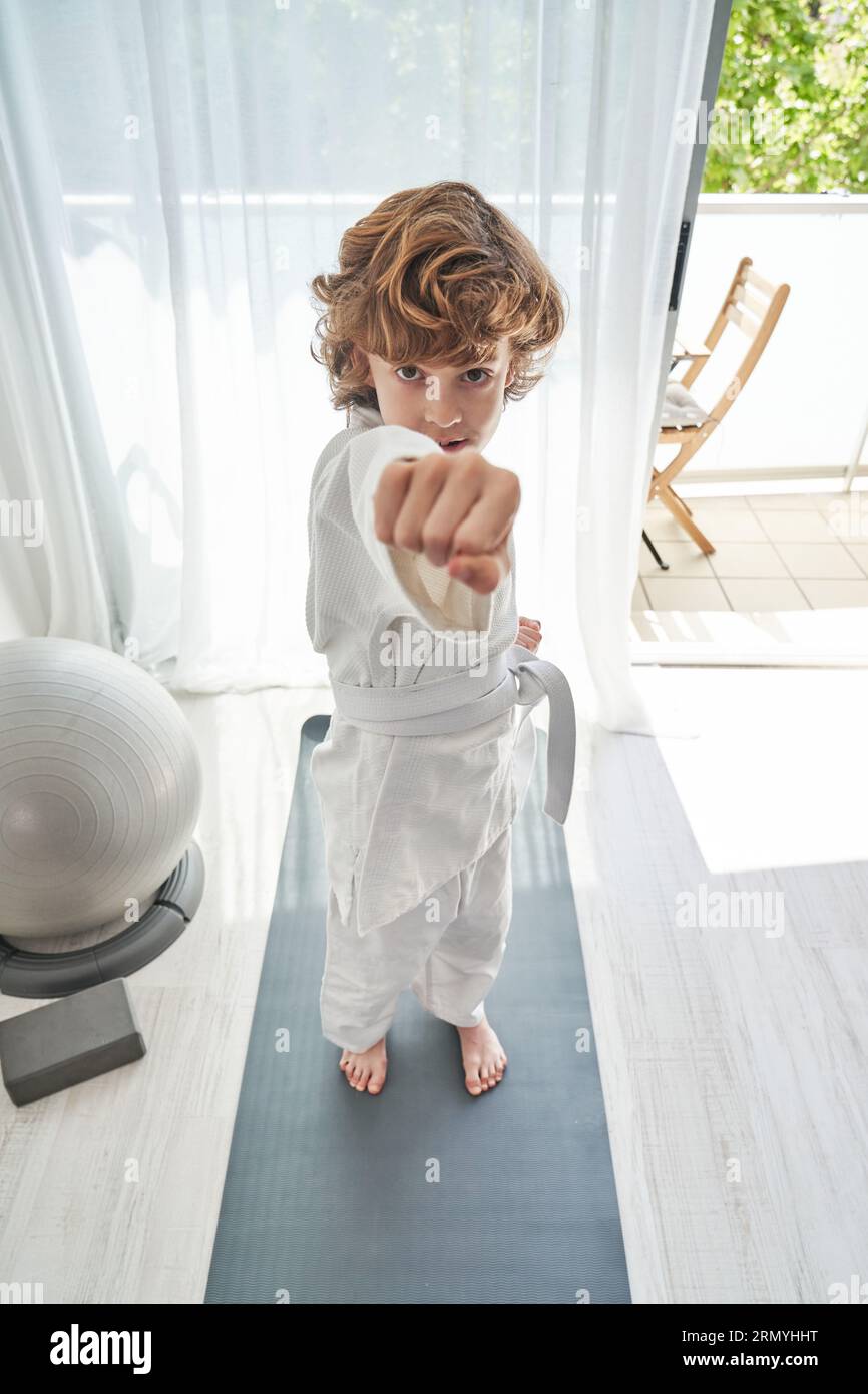 Von oben ein Kind im Kimono mit weißem Gürtel, der auf einer grauen Sportmatte steht und mit geballter Faust auf die Kamera zeigt, während er im Wohnzimmer zu Hause Judo macht Stockfoto