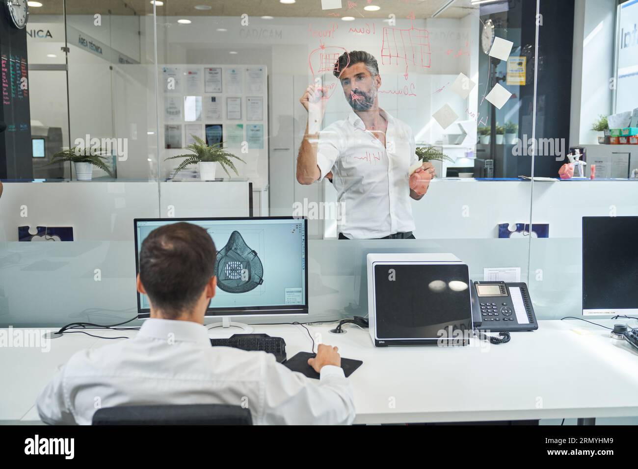 Rückansicht eines anonymen männlichen Arbeiters, der eine Skizze der covid-Maske vorbereitet, während der Kollege ein Schema auf Glaswand im Labor zeichnet Stockfoto