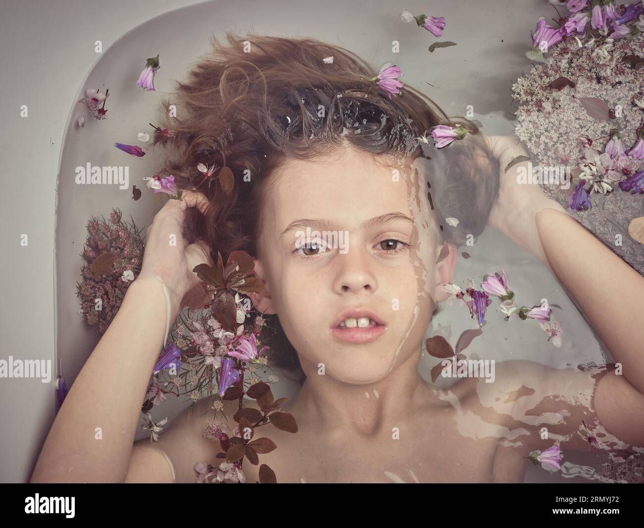 Draufsicht auf den süßen Jungen, der die Haare berührt und die Kamera anschaut, während er in der sauberen Badewanne liegt, mit bunten Blumen im Badezimmer Stockfoto