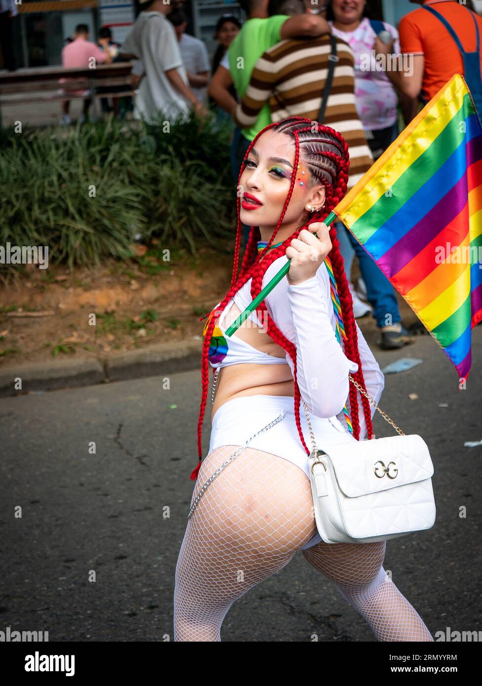 Medellin, Antioquia, Kolumbien - 2. Juli 2023: Junge Frau in sexy weißer Kleidung und roten Zöpfe hält eine Regenbogenfahne und posiert für die Kamera SH Stockfoto