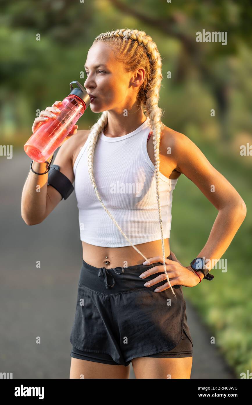 Müde Frau trinkt nach dem abendlichen Workout im Park rote Flasche Wasser. Stockfoto