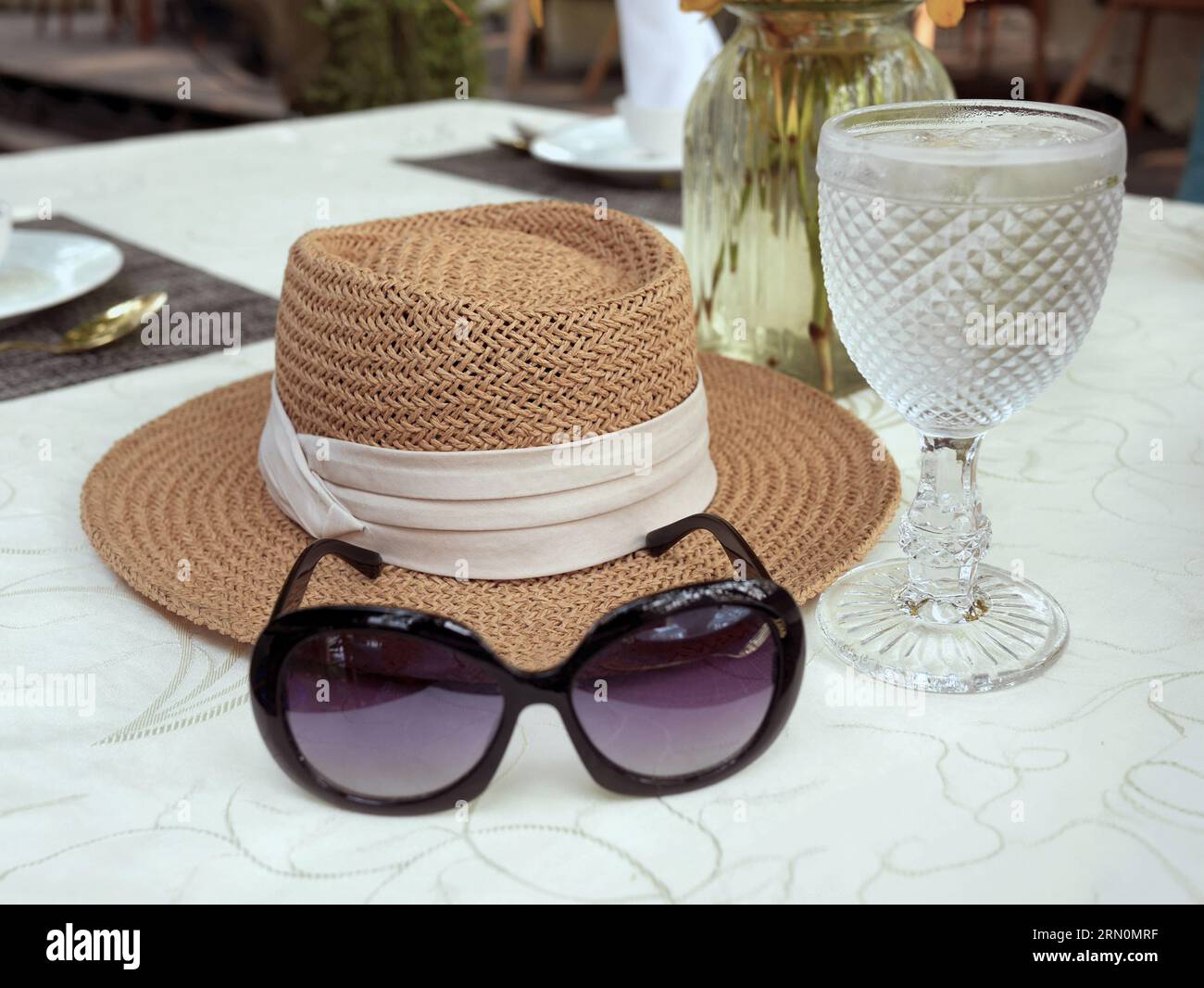 Eine Sonnenbrille und ein Sonnenhut aus gewebtem Stroh, verziert mit beigefarbenem Band auf dem weißen Tischtuch, ein Mittagstisch mit Kristallstielglas und ein va Stockfoto