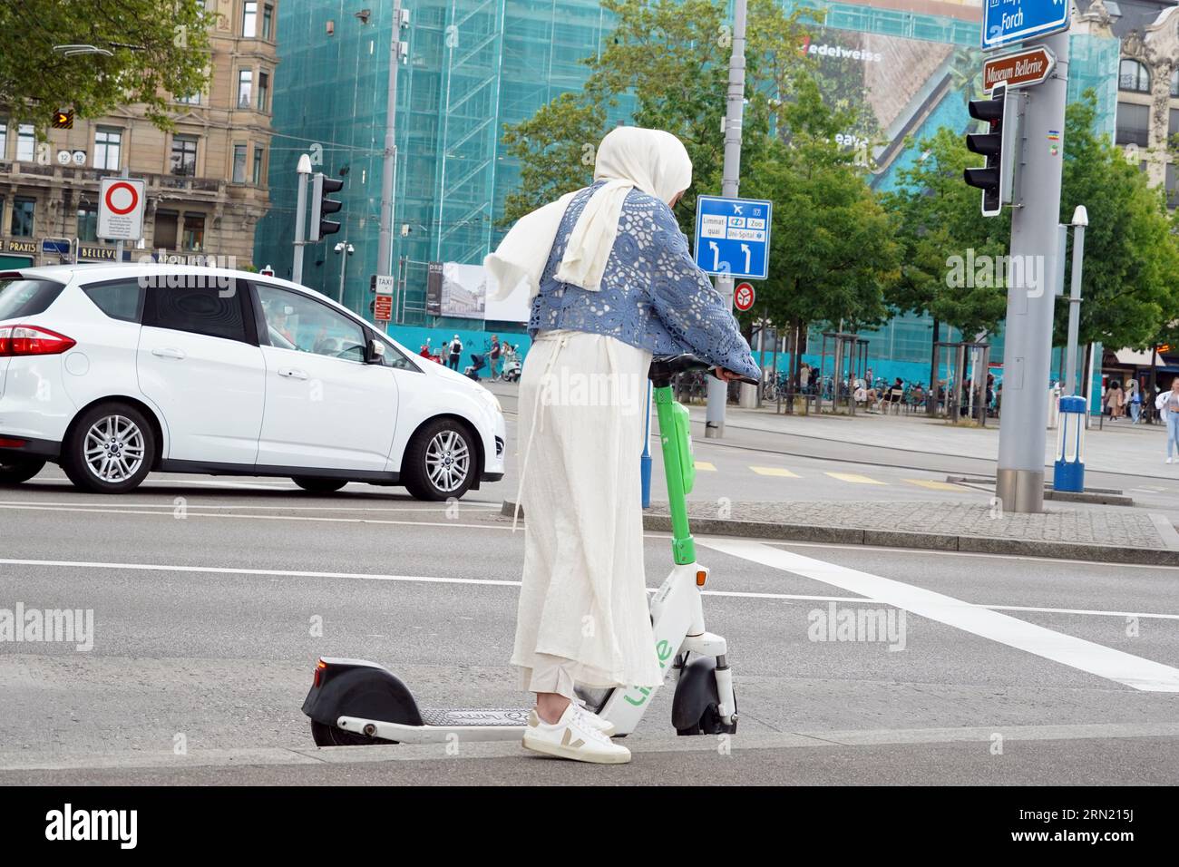 Frau mit weißem Kopftuch, die auf einem Elektroroller an der Kreuzung im Stadtzentrum steht. Stockfoto