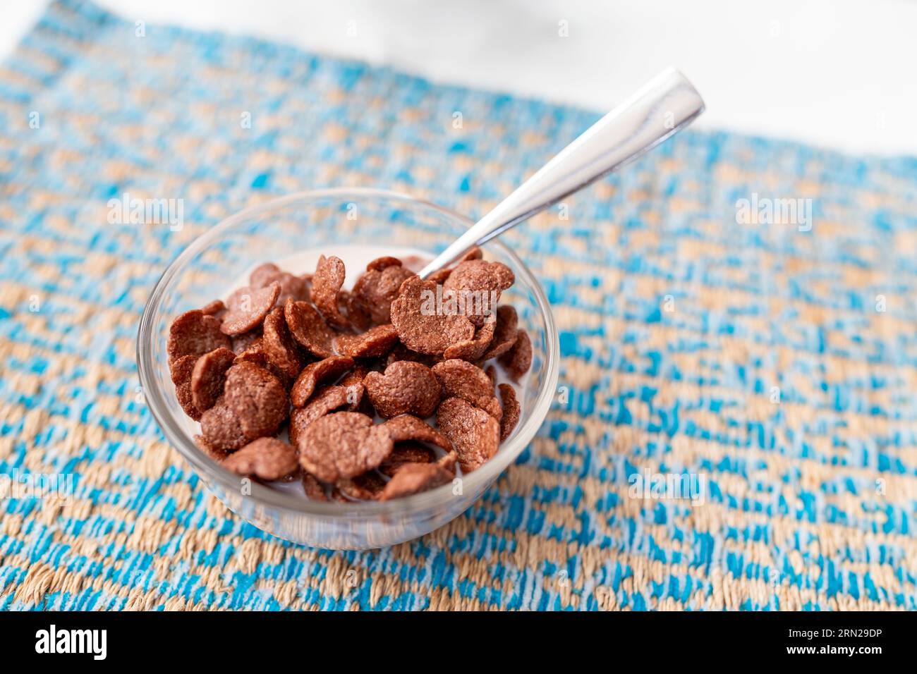 Shot mit flachem Fokus von Frühstückszerealien mit Schokoladengeschmack in einer klaren, runden Glasschale auf einer gemusterten blauen Untertasse, komplett aus Edelstahl Stockfoto