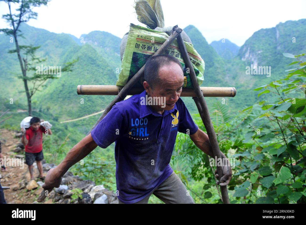 Ein Dorfbewohner trägt Zelte auf seinem Rücken im Dorf Nongwei der Gemeinde Bansheng, südwestchinesische autonome Region Guangxi Zhuang, 18. Juni 2014. Wegen des Mangels an Flachland bauen die meisten Familien in den westlichen Berggebieten von Dahua ihre Häuser auf Hügeln. In den Bezirken Qibainong und Bansheng in Dahua leben Menschen der Miao-Volksgruppe seit etwa tausend Jahren in den Bergen. Sie verlassen sich auf das Pflanzen von Körnern für ihren Lebensunterhalt. Das dreistöckige Stelzengebäude ist im traditionellen Architekturstil der beiden Grafschaften gehalten. Menschen züchten Vieh im unteren Stockwerk, leben auf dem Stockfoto