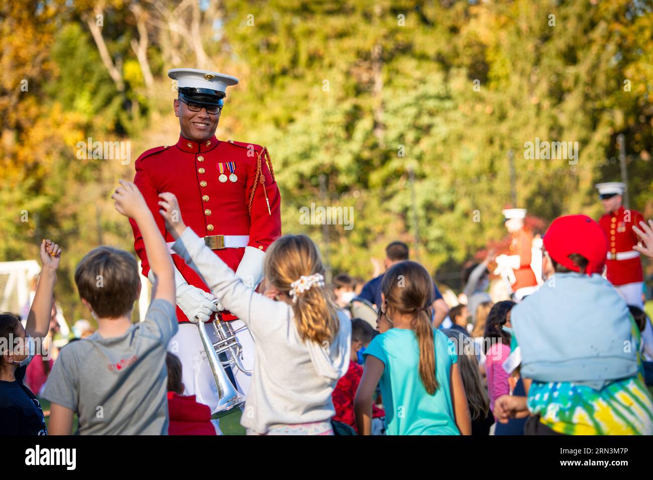 Eigenes Trompeter des Marine Corps Kommandanten mit Schulkindern Arlington Virginia Herbst | ARLINGTON, Virginia, Vereinigte Staaten — Ein Trompeter des US Marine Corps "Commandant's Own" bewegt sich durch eine begeisterte Menge von Grundschulkindern an der Discovery Elementary, Instrument in der Hand, während Kinder die Hand ausstrecken, um ihn zu begrüßen. Das Commandant's Own führt seine Abstammung auf die Feldmusiktraditionen des Marine Corps zurück und dient seit 1934 als das offizielle Trommel- und Trompetenkorps des Corps, das zeremonielle Präzision mit Öffentlichkeitsarbeit verbindet. Stockfoto