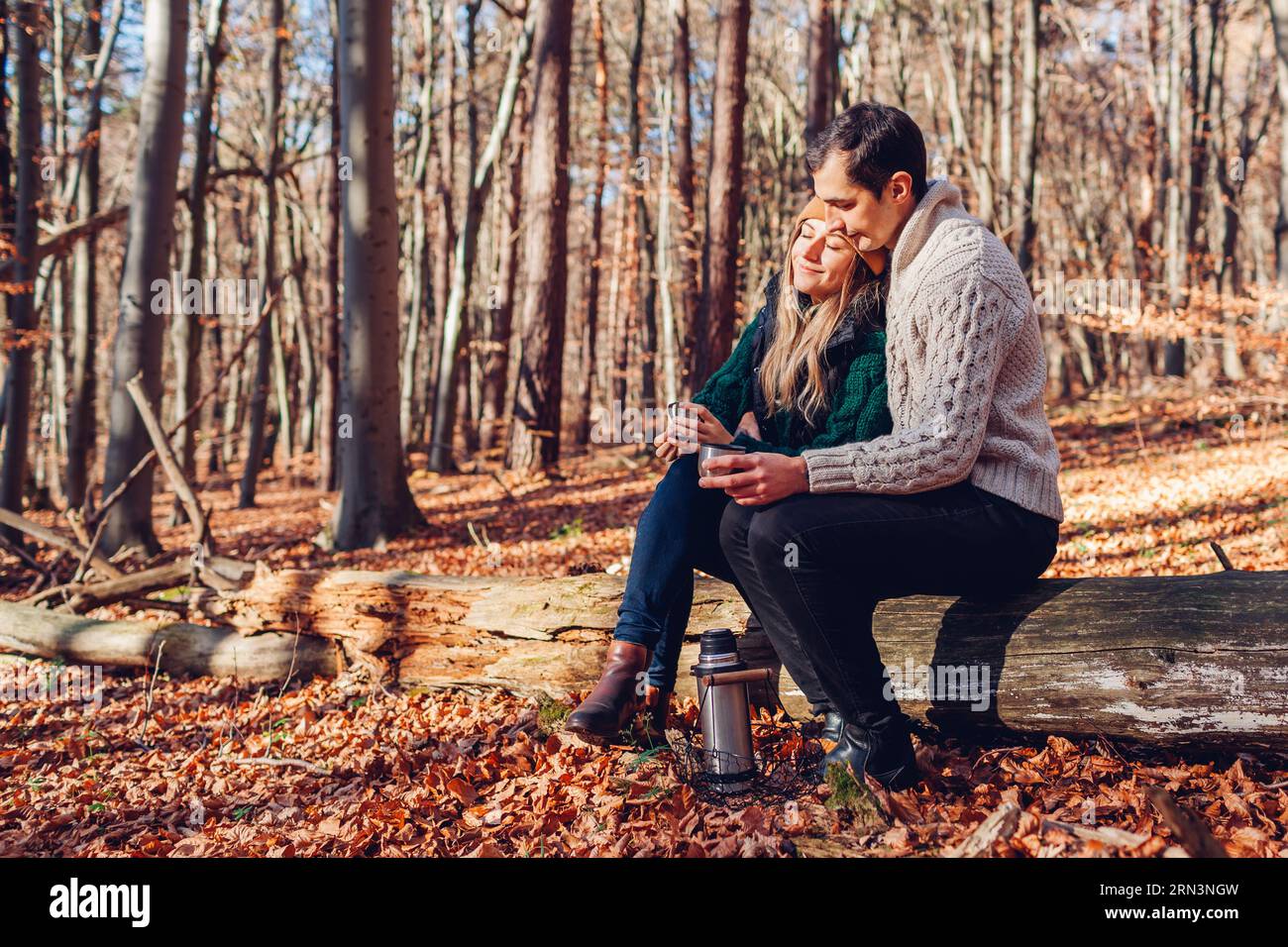 Porträt eines jungen Liebespaares, das Tee trinkt und sich im Herbstwald entspannt. Mann und Frau sitzen auf dem Kofferraum und genießen die herbstliche Landschaft Stockfoto