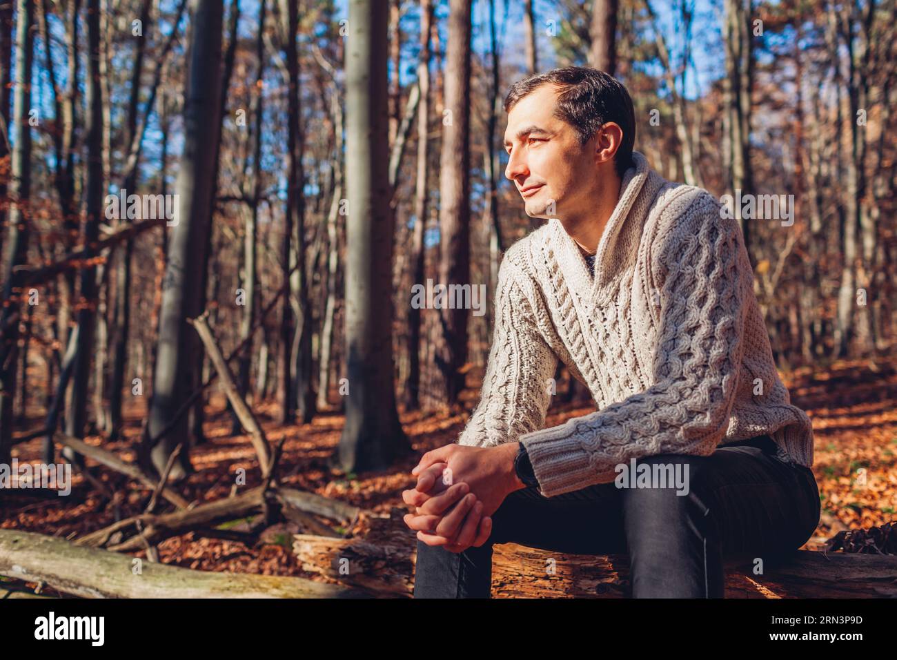 Junger gutaussehender Mann, der sich im Herbstwald entspannt und in Ruhe denkt. Der stilvolle Typ genießt die Herbstlandschaft in einem warmen Pullover. Psychische Gesundheit Stockfoto