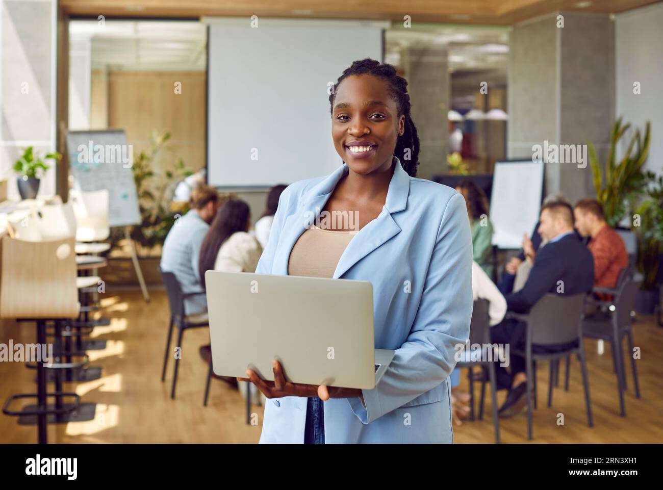 Glückliche, selbstbewusste afroamerikanische Frau, die auf die Kamera schaut, die einen Laptop im Büro hält. Stockfoto