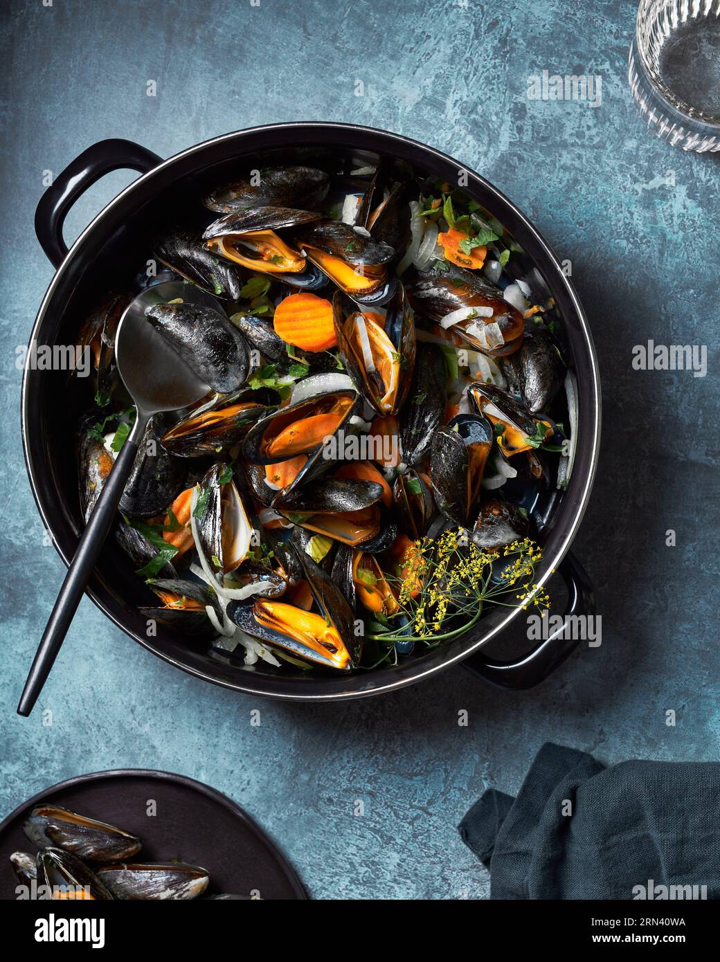 Flachbild von oben von gedämpften und verzehrfertigen Muscheln im schwarzen Topf. Dunkelblauer strukturierter Hintergrund. Glas Wasser, Löffel und Serviette A Requisiten. Stockfoto