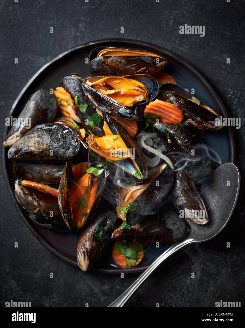 Draufsicht auf eine schwarze Keramikplatte, gefüllt mit gedämpften Muscheln mit Karotten. Da ist Löffel auf dem Teller. Dunkler schwarzer Steinhintergrund. Flachdarstellung vertikal Stockfoto