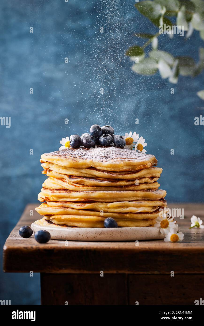 Stapel Pfannkuchen garniert mit Blaubeeren und essbaren Blumen. Es befinden sich Staubpartikel von Zuckerpulver in der Luft. Auf einem alten Holztisch stehen. Stockfoto