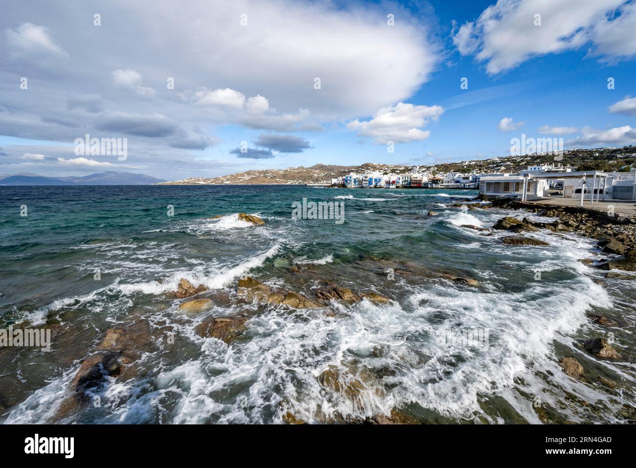 Wellen brechen zwischen den Felsen am Strand, weiße Kykladen auf der Rückseite, Häuser von Little Venice, Mykonos, Griechenland Stockfoto