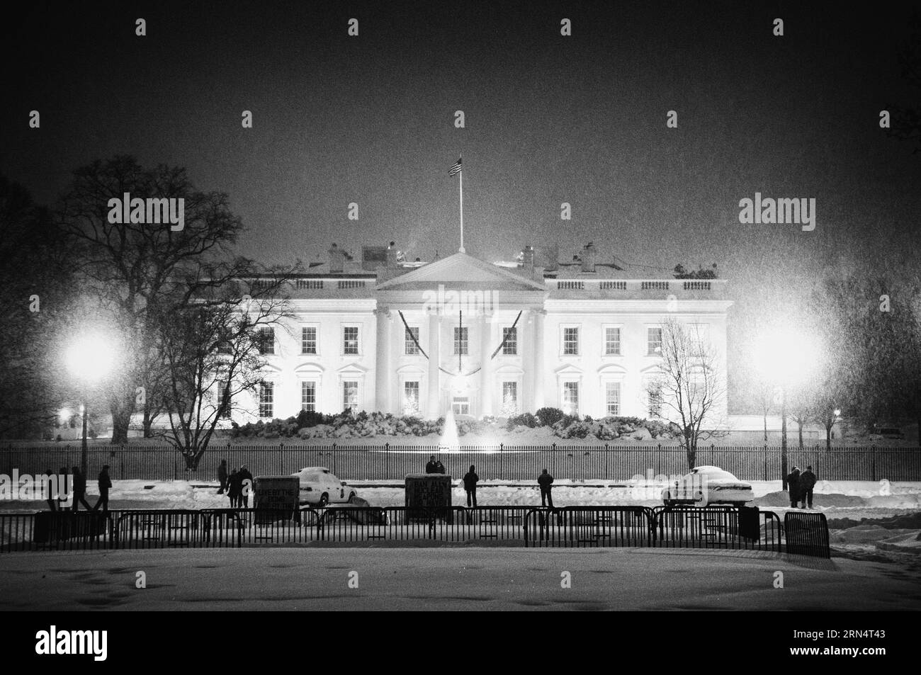 Weißes Haus in Snow, Washington DC — Schwarzweißfoto | WASHINGTON D.C., Vereinigte Staaten – nachts fällt Schnee über das Weiße Haus, während sich Fußgänger und ein Polizeiauto entlang der Pennsylvania Avenue außerhalb des Zauns versammeln. Der North Portico und der Brunnen, der vor dem dunklen Himmel beleuchtet ist, markieren die Residenz, die seit John Adams 1800 für jeden US-Präsidenten erbaut wurde. Stockfoto