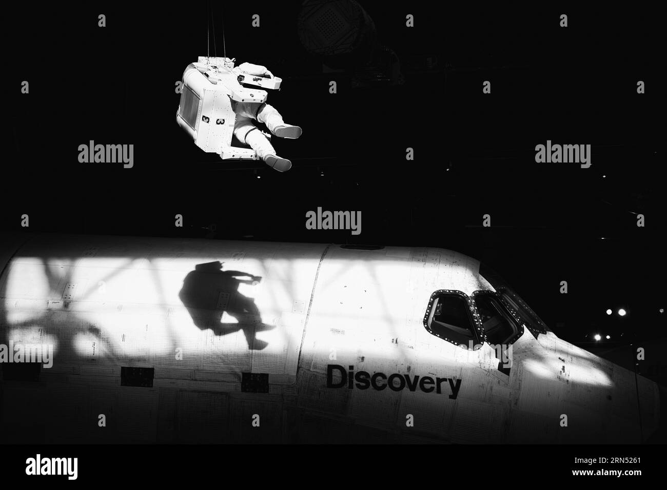 Space Shuttle Discovery and Spacewalk Display, Steven F. Udvar-Hazy Center, Chantilly — Schwarz-weiß-Foto | CHANTILLY, Virginia, USA – Eine Raumfahrt-Astronauten-Ausstellung wirft ihren Schatten über die Nase des Space Shuttle Discovery im Steven F. Udvar-Hazy Center. Discovery flog zwischen 1984 und 2011 39 Missionen, mehr als jeder andere Orbiter, und wird vom Smithsonian als „Fahrzeug der Aufzeichnung“ aufbewahrt. Die Schwarz-weiß-Behandlung betont die Verbrennung der Fliesen aus 39 Wiedereinträgen. Stockfoto
