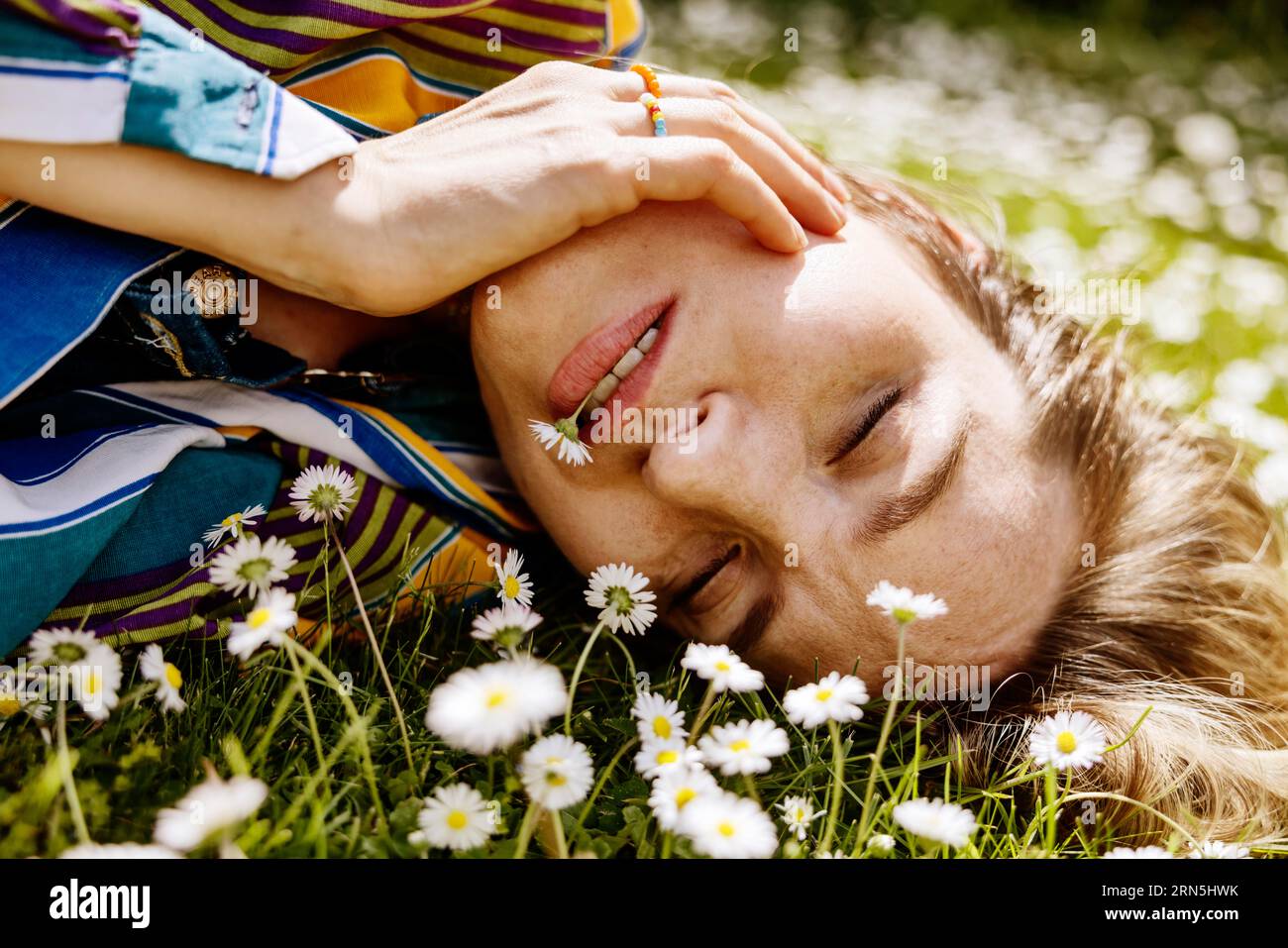 Hübsche junge Frau im Gras zwischen Gänseblümchen mit Gänseblümchen im Mund, Köln, Nordrhein-Westfalen, Deutschland, Wiese, gutes Wetter Stockfoto
