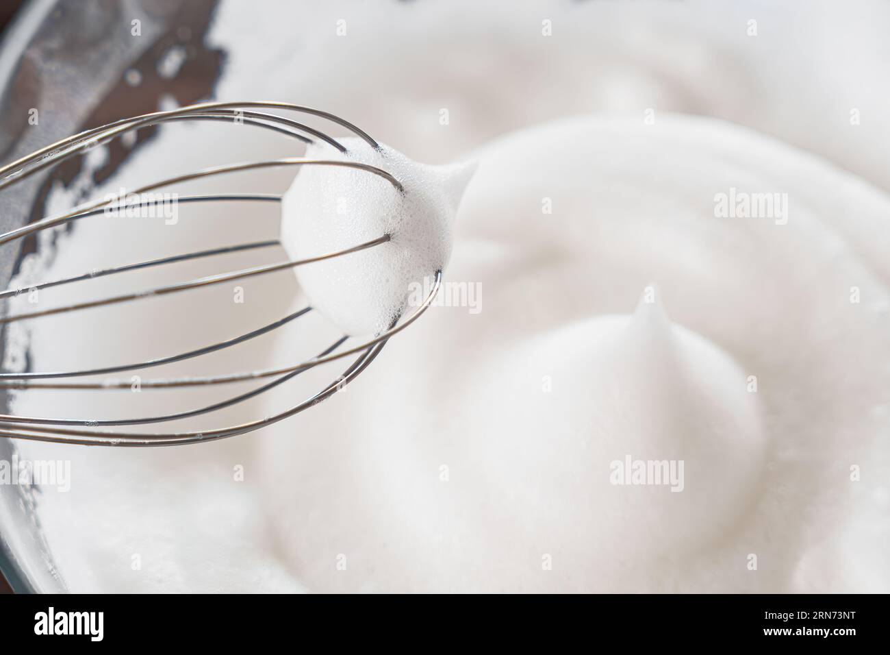 Nahaufnahme von einem Schneebesen und geschlagenem Eiweiß zu steifen Gipfeln, ein Schritt bei der Zubereitung von Frühstücksteig oder zarten Pfannkuchen. Weißer geschlagener Schaum aus Ei W Stockfoto