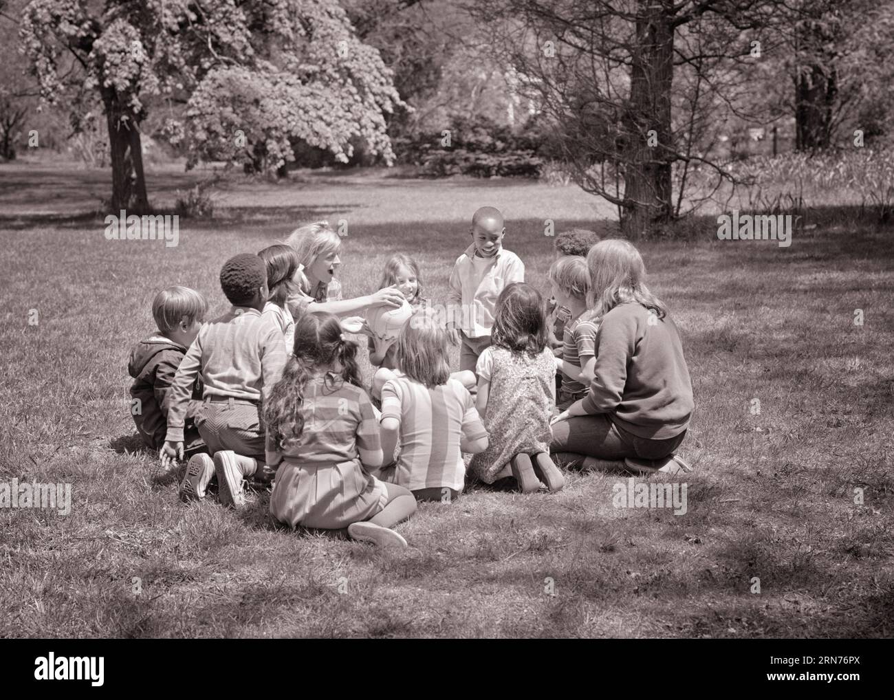 1960ER JAHRE ERWACHSENE LEHRERIN, DIE EINEN BALL AN EINE ETHNISCH GEMISCHTE GRUPPE VON KINDERN VERSCHIEDENEN ALTERS IM KREIS AUF DEM ÜBERGIBT PARK GRASS - J12340 HAR001 HARS GESUNDHEIT KOPIE RAUM FREUNDSCHAFT HALBE LÄNGE DAMEN PERSONEN INSPIRATION HANDING MÄNNCHEN B&W GEMISCHTES ALTER GLÜCK HIGH-ANGLE ABENTEUER ENTDECKUNG FREIZEIT AFROAMERIKANER AFROAMERIKANER AUFREGUNG INSTRUKTOR BLACK ETHNICITY BERUFE KONZEPTIONELLE ERZIEHER VERSCHIEDENE NACH DER SCHULE ZUSAMMENARBEIT PÄDAGOGISCHE ERZIEHER ETHNISCHE WACHSTUMSLEHRER JUGENDLICHE SCHULE LEHRT ZWEISAMKEIT JUNGE ERWACHSENE FRAU SCHWARZ-WEISS KAUKASISCHE ETHNIE HAR001 ALTMODISCH Stockfoto