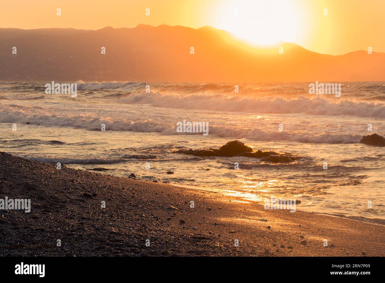 Wellen an einem idyllischen Strand in Griechenland. Sommertag mit rauem Meer. Im Hintergrund untergeht die Sonne hinter den Bergen Stockfoto