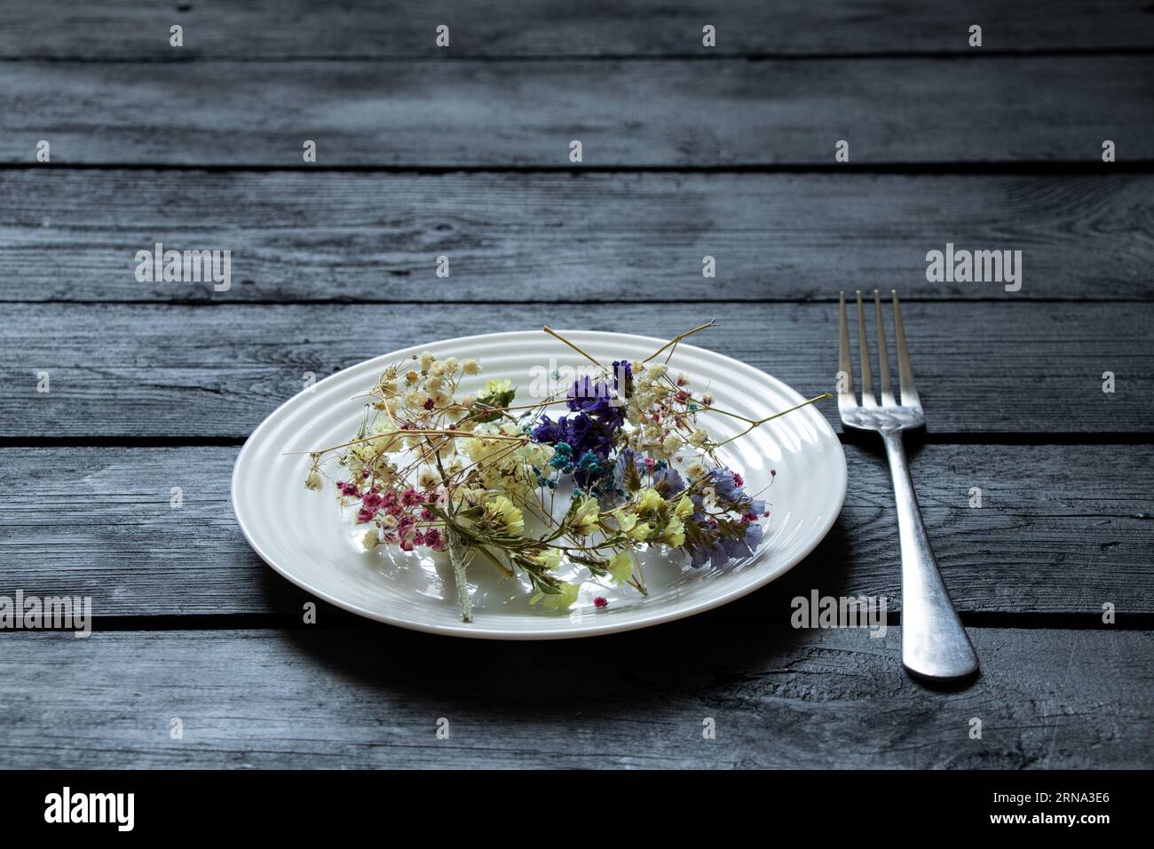 Weißer Teller mit verschiedenen getrockneten Blumen auf einem schwarzen Holzbrett und einer Gabel in der Nähe, Blumen und Essen, Frühling Stockfoto