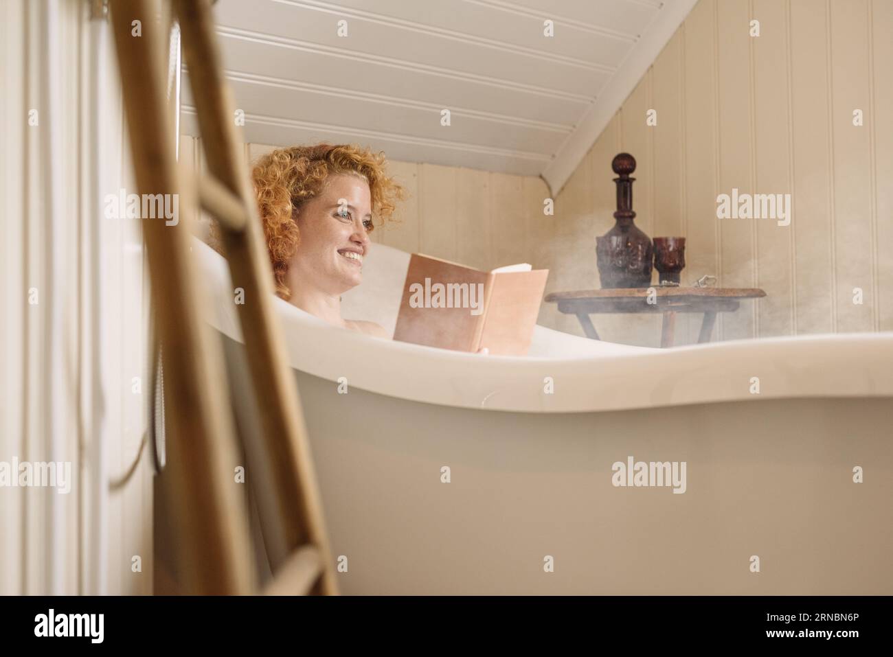Fröhliche junge Frau, die in der Badewanne liegt und bei Tageslicht Buch liest Stockfoto