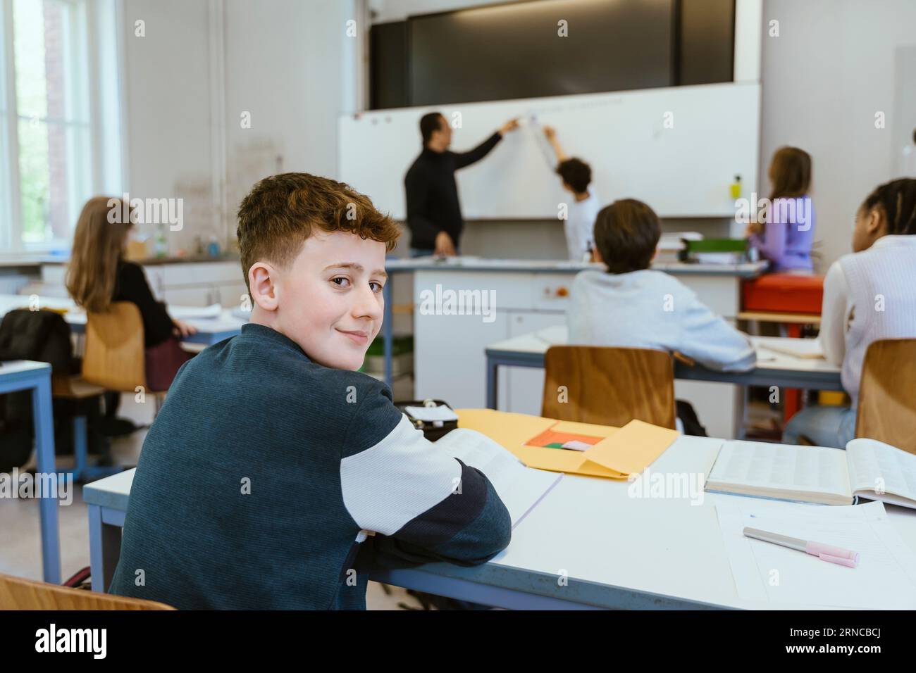 Porträt eines lächelnden Jungen, der über die Schulter schaut, während er im Klassenzimmer auf dem Schreibtisch sitzt Stockfoto