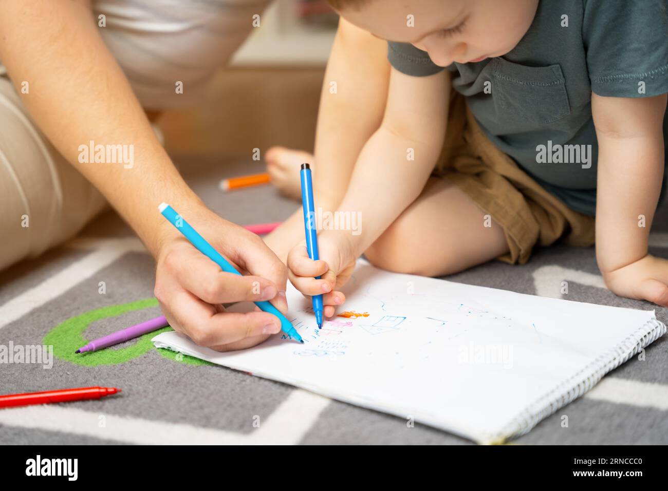 Ein wunderschöner kleiner kleiner Junge von zwei oder drei Jahren mit Vater zeichnet mit Markern im Album im Kinderzimmer zu Hause auf dem Boden. Stockfoto